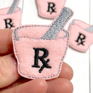 May include: Four pink felt patches with an embroidered mortar and pestle and the letter "R" with a cross through it. The patches are approximately 2 inches in diameter.