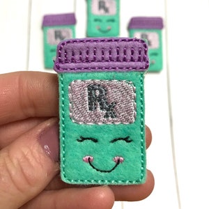 May include: A set of four green felt pill bottle ornaments with purple lids. Each bottle has a smiling face and the letter "R" on the front.
