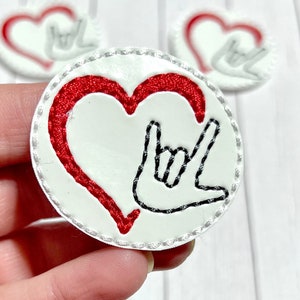 May include: A white heart-shaped patch with a red heart inside and a black embroidered hand sign for "I love you" in American Sign Language.