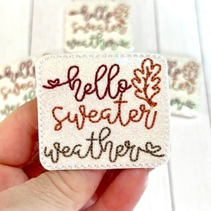 May include: White felt patch with the text "hello sweater weather" in brown and orange embroidery. A small brown leaf is embroidered next to the text.