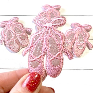 May include: Three pink glitter ballet shoe appliques with a bow on each pair. The appliques are on a white wooden surface.