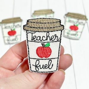 May include: White coffee cup with a brown lid and a red apple with a green leaf on the front. The cup says "Teacher Fuel" in black cursive writing.