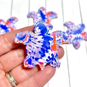 May include: Tie-dye sea turtle-shaped embellishments in shades of blue, orange, and white. Each turtle has a silver glitter outline. The image shows several of these decorative elements.