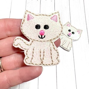 May include: Two white cat-shaped fabric patches with a subtle glitter finish. Each patch features embroidered details including black circular eyes, a small pink nose, a stitched mouth, and pink inner ears, all outlined with tan decorative stitching.