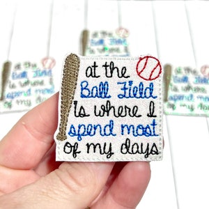 May include: White baseball-themed patch with the text "at the Ball Field is where I spend most of my days." The patch features a baseball bat, a baseball, and blue and black embroidered lettering. The patch is held in a hand.