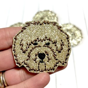 May include: A gold glitter iron-on patch with a brown outline of a dog's face. The dog has black eyes and a black nose.