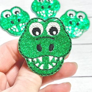 May include: Four green glitter alligator head patches with black outlines, white teeth, and black eyes. One patch is held in a hand, with three others in the background. The patches are on a white wooden surface.