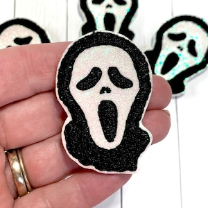 May include: Close-up of several glittery white and black Scream mask-shaped embellishments. The masks have a black outline and a white face with an open mouth. The glitter adds a sparkling effect. These are likely for crafting or Halloween decor.