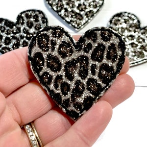 May include: Close-up of several heart-shaped appliques with a leopard print design. The appliques are black and silver glitter, with a black border. The hearts are on a white background.