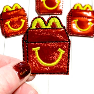 May include: Four red and yellow iron-on patches shaped like a McDonald's fast food box with the iconic golden arches and a smiling face. The patches have a glittery finish.