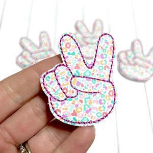 May include: Peace sign patches with a colourful leopard print pattern. The patches are white with a pattern of pink, orange, blue, and yellow spots. Each patch has a purple stitched outline.