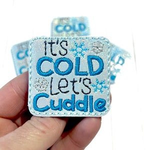 May include: A blue and white embroidered patch with the text "It's Cold Let's Cuddle" and snowflake designs.