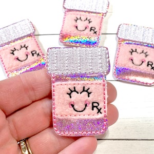 May include: Four pink and silver glitter pill bottle patches with a smiling face and the letters "Rx" embroidered on them.