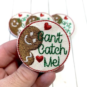 May include: A white, round, felt patch with a gingerbread man design and the text "Can't Catch Me!" in green and red embroidery. There are two red hearts on the patch.