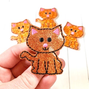 May include: A set of four orange glitter cat-shaped patches. Each patch features a bright orange glitter finish, black embroidered outlines, pink inner ears, a pink nose, and two black circular eyes. The cat is depicted in a sitting position.