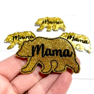 May include: Gold glitter bear-shaped embellishments with the word "Mama" in black. The bear has a black outline. Several similar embellishments are visible in the background.
