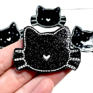 May include: Black glitter cat-shaped patches with white stitched details and a small white heart. The patches have a sparkly texture and are arranged in a group. The cat patches are a craft supply.