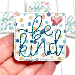 May include: Square patch with the words "be kind" embroidered in teal thread on a white background with colorful polka dots. A small red heart is embroidered above the "d". The patch has a stitched border.