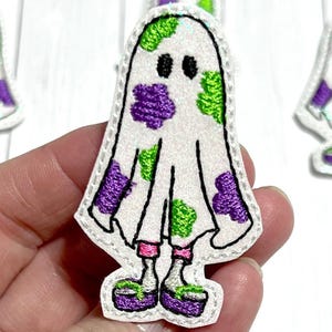 May include: White ghost with purple and green splotches, wearing purple and green shoes. The ghost is embroidered on a white fabric.