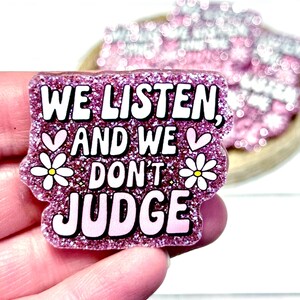 May include: A pink glitter badge with the words "WE LISTEN, AND WE DON'T JUDGE" in white, with heart and daisy accents. The badge is held in a hand, with other similar badges in the background.