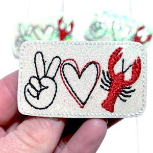 May include: A rectangular patch with the words "Peace, Love, Crawfish" embroidered on a glittery, off-white background. The design includes a peace sign, a heart, and a red crawfish.