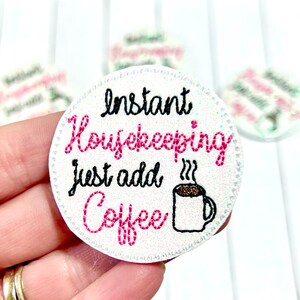 May include: A white, round patch with the text "Instant Housekeeping just add Coffee" in black and pink script. A steaming coffee cup graphic is also included. The patch has a glittery border and is likely an accessory.