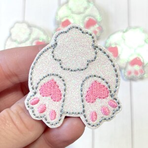 May include: A glittery white applique in the shape of a bunny's rear end, featuring pink paw pads and a gray stitched outline. Several similar appliques are visible in the background, suggesting a craft or DIY project.