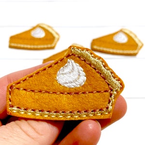 May include: A felt pumpkin pie slice with a white whipped cream topping. The pie slice is decorated with a gold embroidered edge.