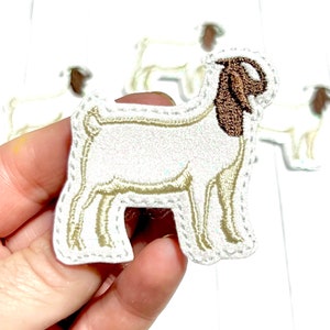 May include: White embroidered goat applique with brown ears and a white background. The applique is held in a hand.