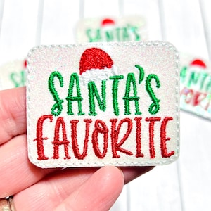 May include: A rectangular patch with the text "SANTA'S FAVORITE" in green and red embroidery, with a Santa hat graphic. The patch has a white background with glitter.