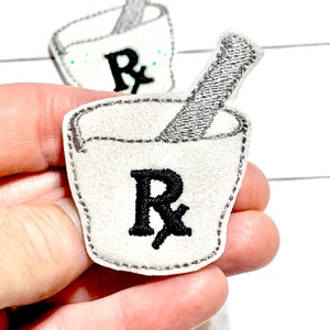 May include: Two white felt patches with an embroidered mortar and pestle and the letter "R" in black. The patches are approximately 2 inches in diameter.