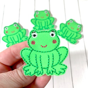 May include: Three green felt frog appliques with embroidered details. The frogs have black eyes, pink cheeks, and a pink smile. The frogs are sitting on a white surface.
