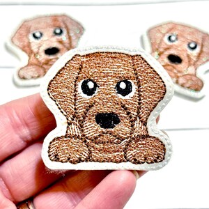 May include: Embroidered patch of a brown puppy with large black eyes and a black nose. The patch has a white border and is designed to look like a friendly dog. Two more patches are visible in the background.