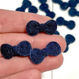 May include: A close-up of a hand holding multiple denim blue fabric bows. The bows are small and have a textured surface.