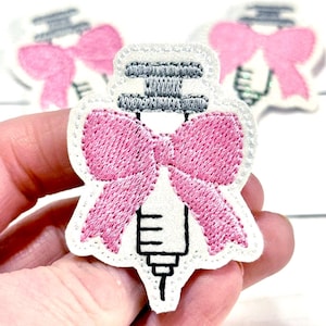 May include: Embroidered patch with a syringe design and a pink bow. The patch has a white border and a grey syringe plunger. The bow is a vibrant pink, adding a decorative touch to the medical-themed patch.
