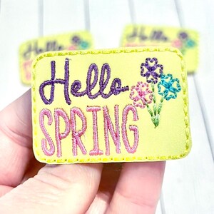 May include: A yellow embroidered patch with the words "Hello SPRING" in purple and pink thread. The patch features embroidered flowers in pink, purple, and blue. The patch is rectangular with a stitched border.