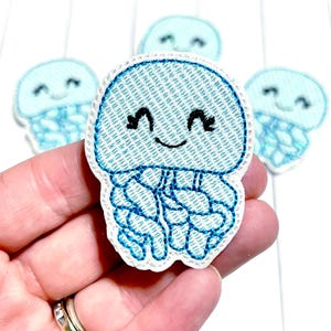 May include: A collection of light blue jellyfish patches with embroidered details. Each patch features a smiling face and flowing tentacles. The patches are made of fabric and have a white border. The patches are suitable for sewing or ironing onto clothing or accessories.