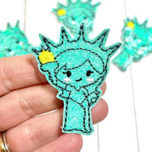May include: A collection of teal felt Statue of Liberty patches with black stitching and yellow accents. The patches feature a cartoon-like design with smiling faces and are held in a hand.