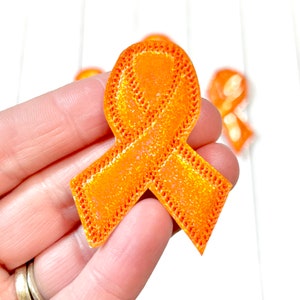 May include: An orange awareness ribbon with a glitter finish and a stitched edge. The ribbon is held in a hand, with other ribbons visible in the background against a white surface.