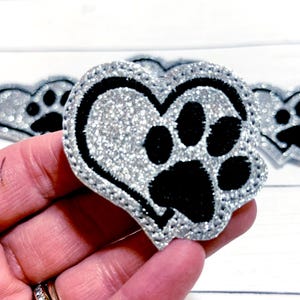May include: A silver glitter heart-shaped patch with a black paw print design. The patch has a black border and is held in a hand. The background is a white wooden surface.