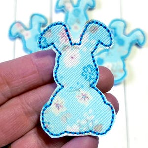 May include: A light blue bunny-shaped applique with a floral pattern and a blue stitched outline. Several similar appliques are visible in the background, suggesting a craft or DIY project. The applique is held in a hand.