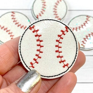 Peut inclure: Un accessoire rond sur le thème du baseball, blanc avec des coutures rouges et un bord noir. L'accessoire a une texture pailletée. Plusieurs articles similaires sont visibles en arrière-plan.