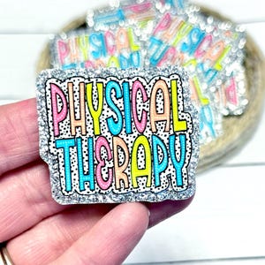May include: A glittery badge with the words "PHYSICAL THERAPY" in colorful bubble letters. The letters are outlined in black and set against a white background with black polka dots. The badge has a silver glitter border.