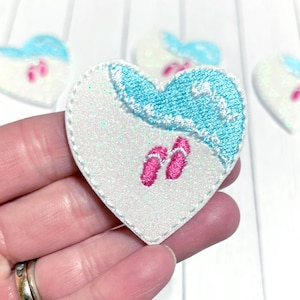 May include: Heart-shaped applique with a beach theme. The design features a white glittery base, a turquoise wave, and pink flip-flops. Additional heart-shaped appliques are visible in the background. The applique is held in a hand.