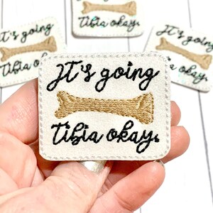 May include: White felt patch with black embroidery that reads "It's going tibia okay" with a brown bone graphic.