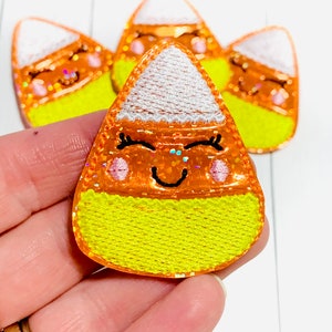 May include: Four embroidered iron-on patches shaped like candy corn with a smiling face. The patches are yellow, orange, and white with glitter accents.