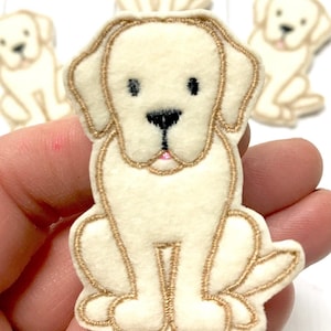 May include: A cream-colored felt patch shaped like a sitting Labrador retriever puppy. The design features embroidered gold-toned outlines, black thread for the eyes and nose, and a small pink accent for the mouth, creating a detailed canine figure.