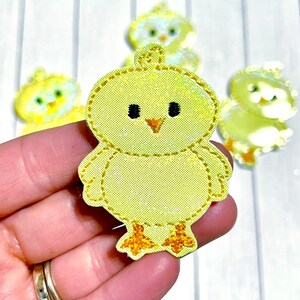 May include: Yellow chick applique with black eyes and an orange beak. The chick is made of fabric and has a stitched outline. Several other chick appliques are in the background. The appliques are likely for crafts or Easter decorations.