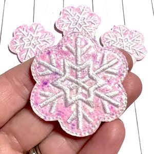 May include: Four pink glitter snowflake embellishments with white embroidery. The snowflakes are approximately 2 inches in diameter.