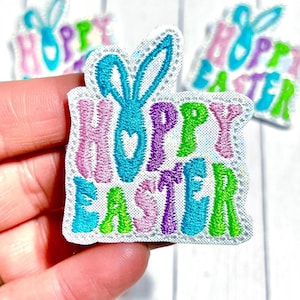 May include: Embroidered patch with the words "Happy Easter" in colourful letters. The patch features a bunny ear design in turquoise, purple, green, and pink. The patch is white with a decorative border.